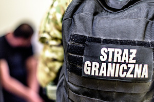 Na pierwszym planie napis Straż Graniczna. W oddali siedzi zatrzymany. Na pierwszym planie napis Straż Graniczna. W oddali siedzi zatrzymany.