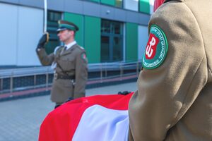 Funkcjonariuszka trzyma na rękach Flagę Polski. W tle dowódca prezentuje broń. Funkcjonariuszka trzyma na rękach Flagę Polski. W tle dowódca prezentuje broń.