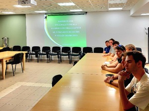 Uczniowie siedzą przy stole. W tle ekran z wyświetlaną prezentacją. Uczniowie siedzą przy stole. W tle ekran z wyświetlaną prezentacją.
