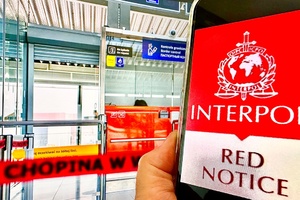 Czerwona Nota Interpolu wyświetlona na telefonie trzymanym w ręku. W tle kabina kontrolerska. Czerwona Nota Interpolu wyświetlona na telefonie trzymanym w ręku. W tle kabina kontrolerska.