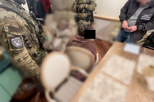 Zatrzymany siedzi na krześle. Obok niego stoją funkcjonariusze. Zatrzymany siedzi na krześle. Obok niego stoją funkcjonariusze.